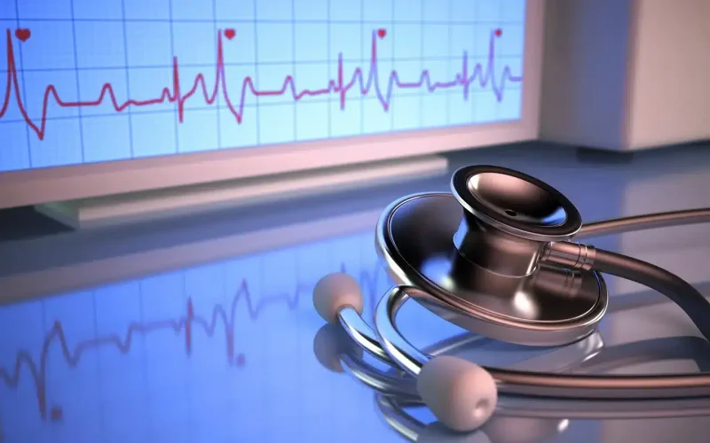 A stethoscope lies on a reflective surface in front of a monitor displaying a heart rate electrocardiogram (ECG) graph.