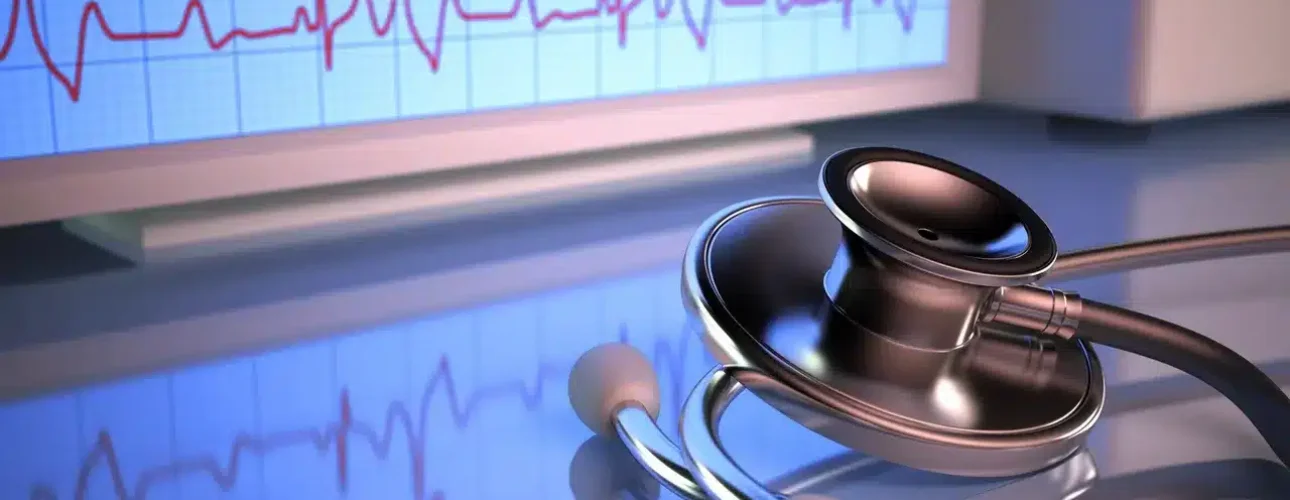 A stethoscope lies on a reflective surface in front of a monitor displaying a heart rate electrocardiogram (ECG) graph.
