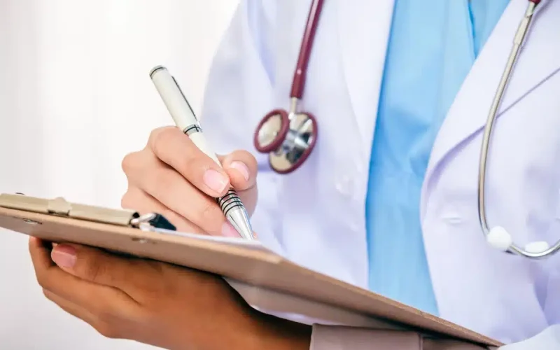 A person wearing a white medical coat and stethoscope writes on a clipboard with a pen.