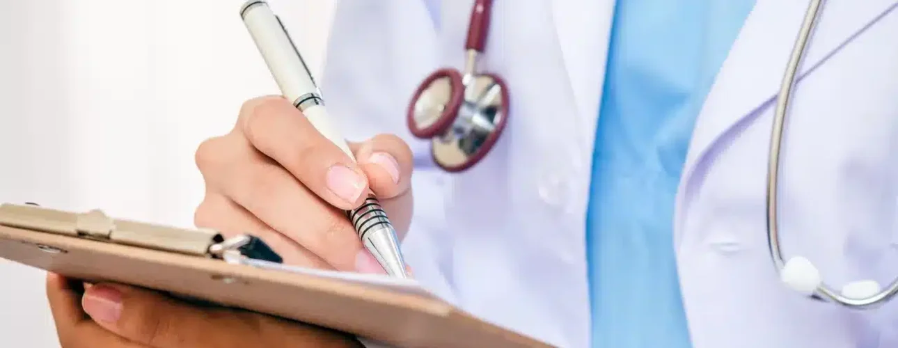 A person wearing a white medical coat and stethoscope writes on a clipboard with a pen.